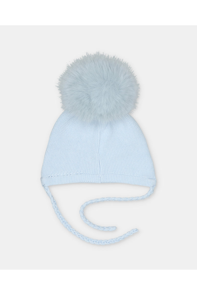 A light blue knit baby beanie hat with a fluffy pom-pom on top and braided ear ties, shown against a plain light gray background.