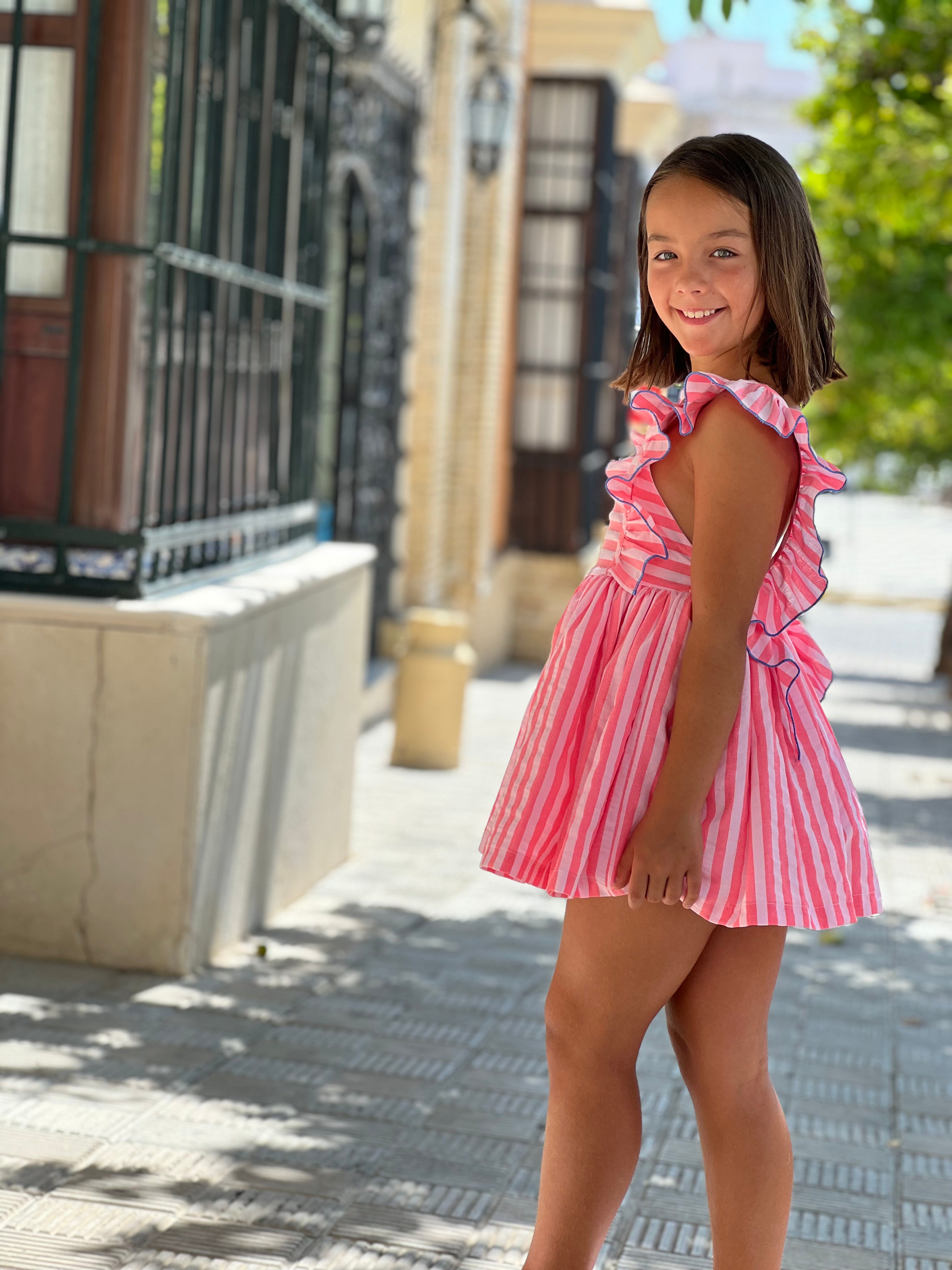 Young girl in a La Peppa pink and white striped dress standing on a sidewalk.