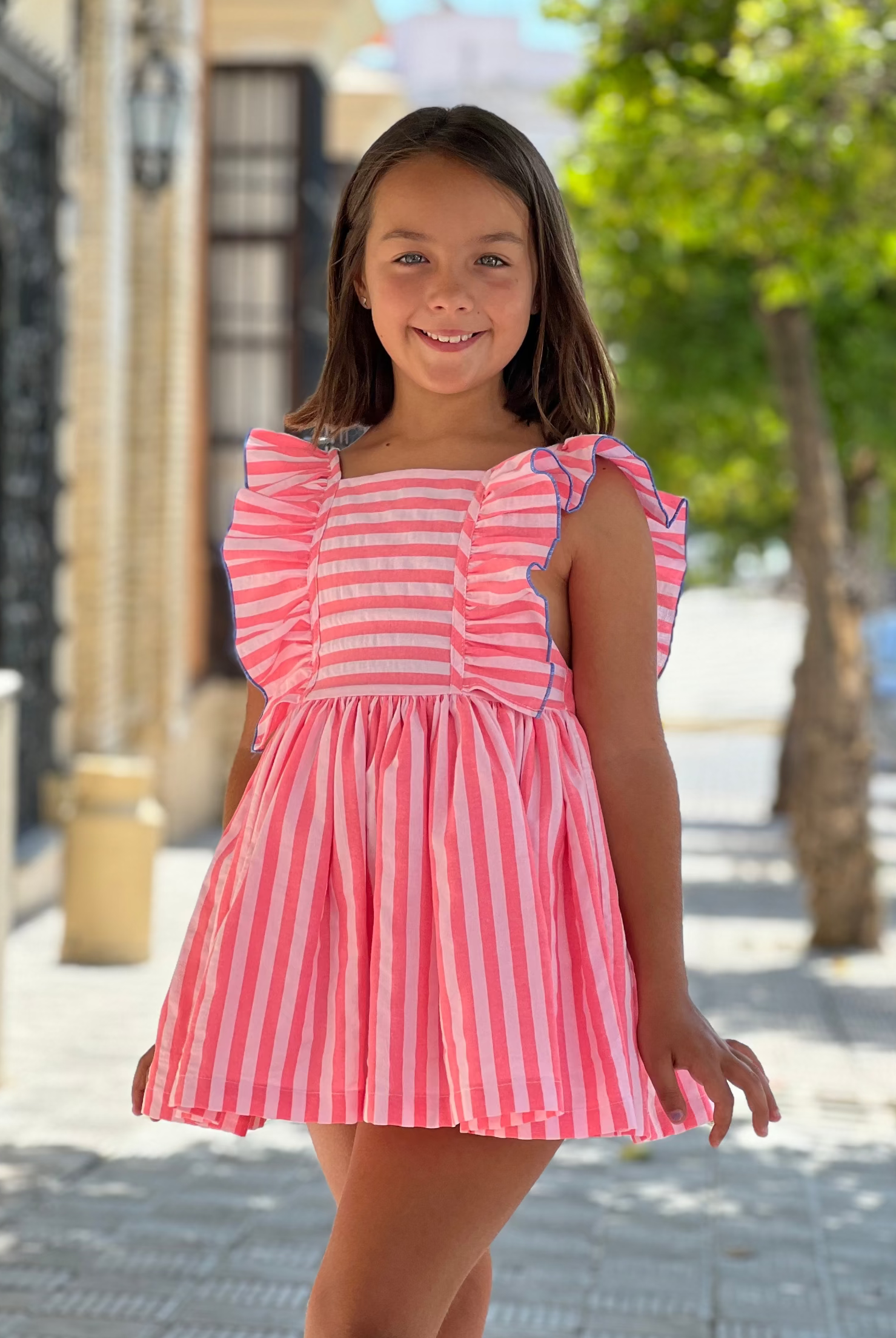Young girl in a pink and white striped dress standing outdoors on a sunny day.
