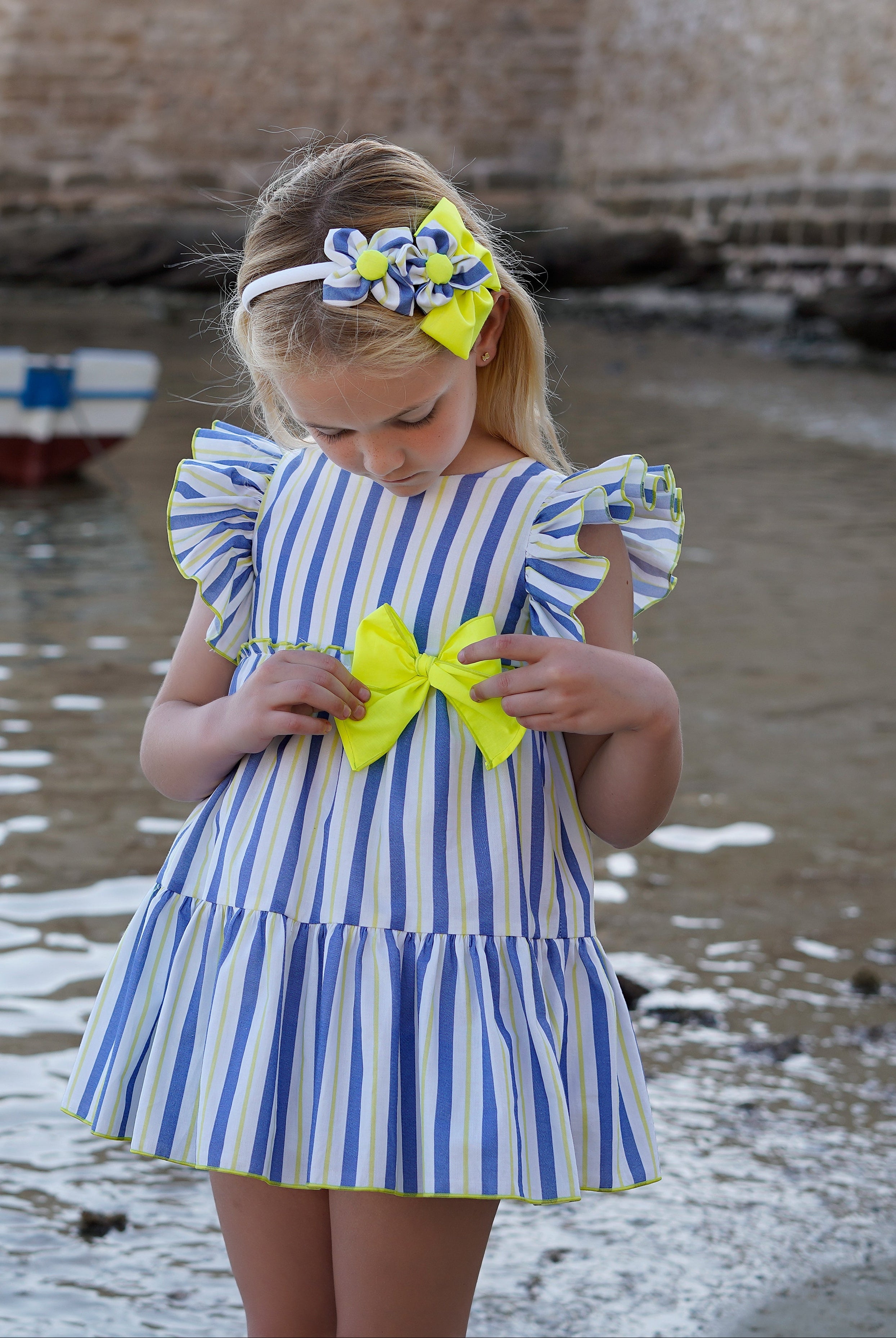 A young girl stands on a sandy shore by the water, wearing a blue and white vertically striped tiered dress with ruffled sleeves and a large yellow bow at the waist. She also wears a matching yellow and blue flower headband.