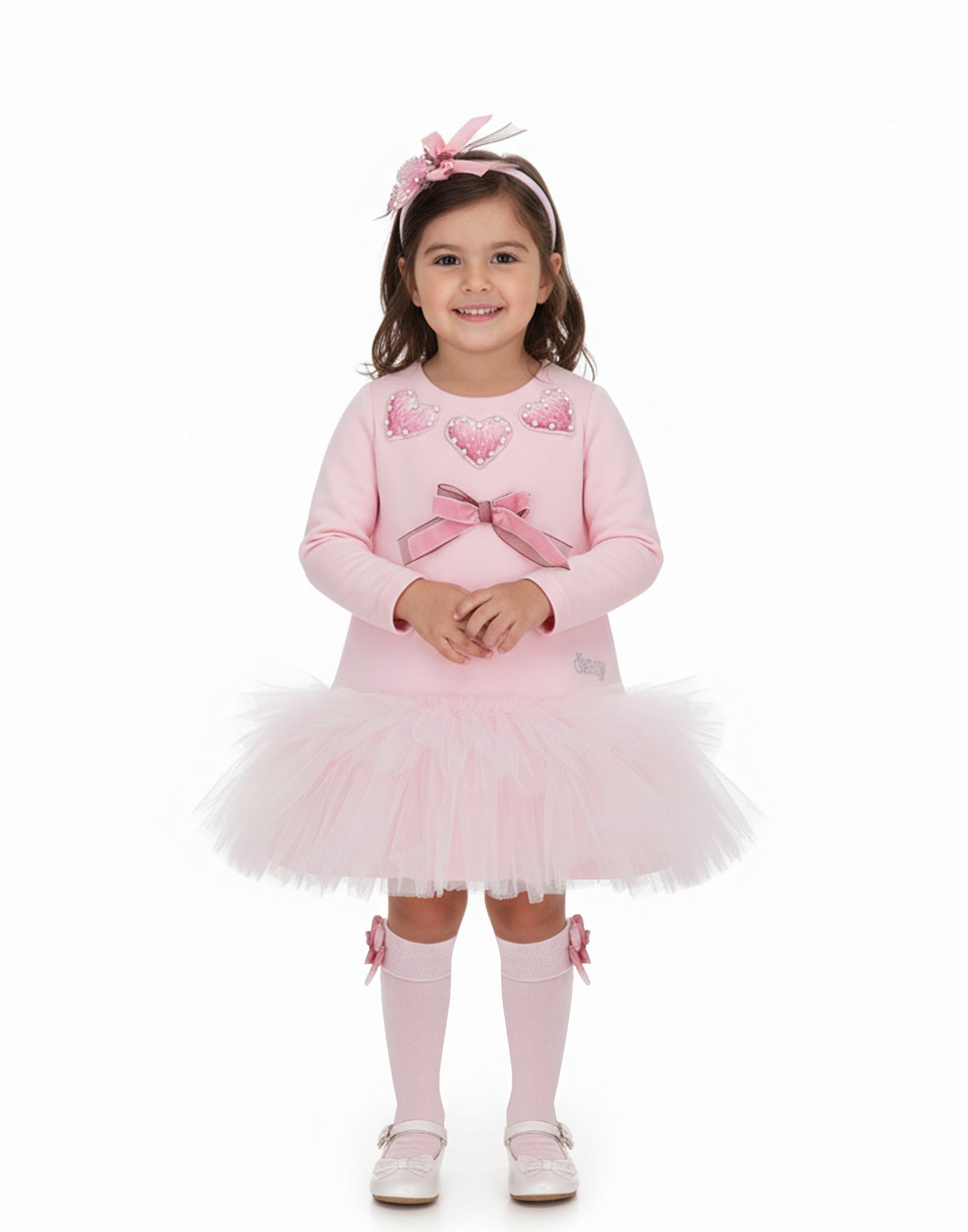 A young girl stands against a plain white studio background wearing a light pink long-sleeve dress with a tulle tutu skirt, pink bow details, matching pink socks, and a pink bow headband.