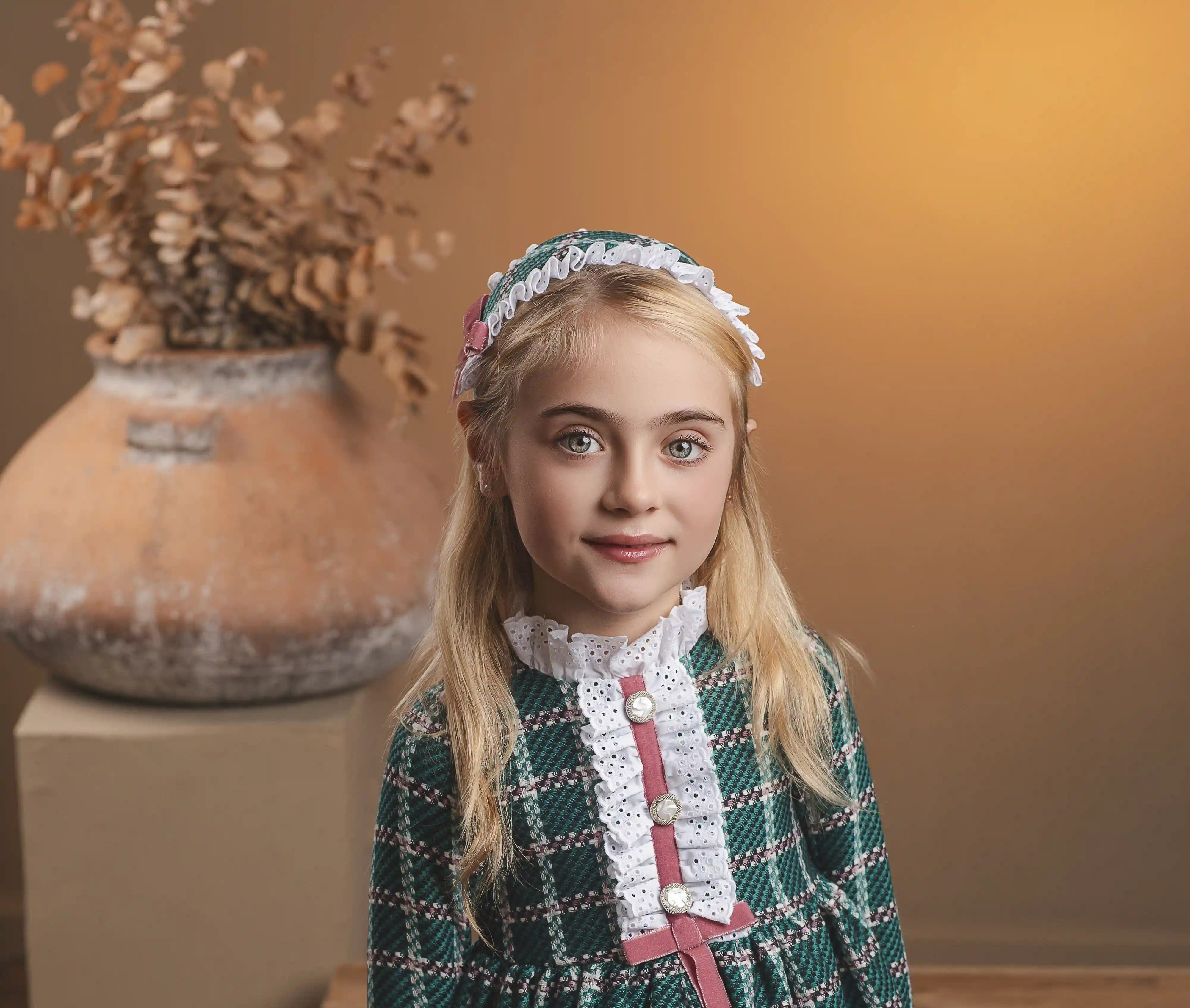 A young girl with blonde hair stands wearing a green and white plaid dress with a pink bow and white lace collar, paired with a matching headband. She is posed in a warm, rustic indoor setting with a wooden bench and a large terracotta vase in the background.