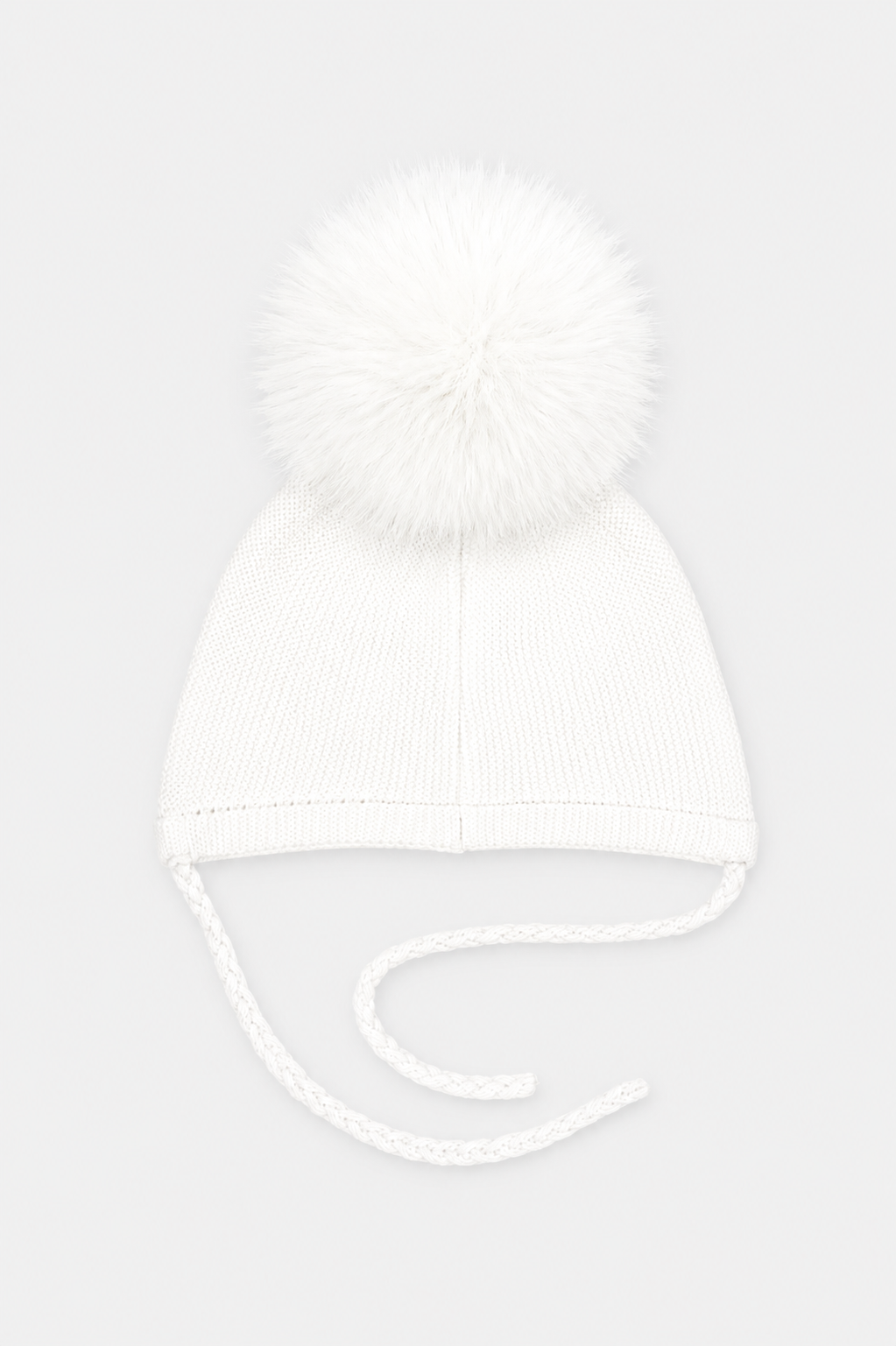 A white knit baby beanie hat featuring a large fluffy pom-pom on top and matching white ear tie strings, shown against a light gray background.