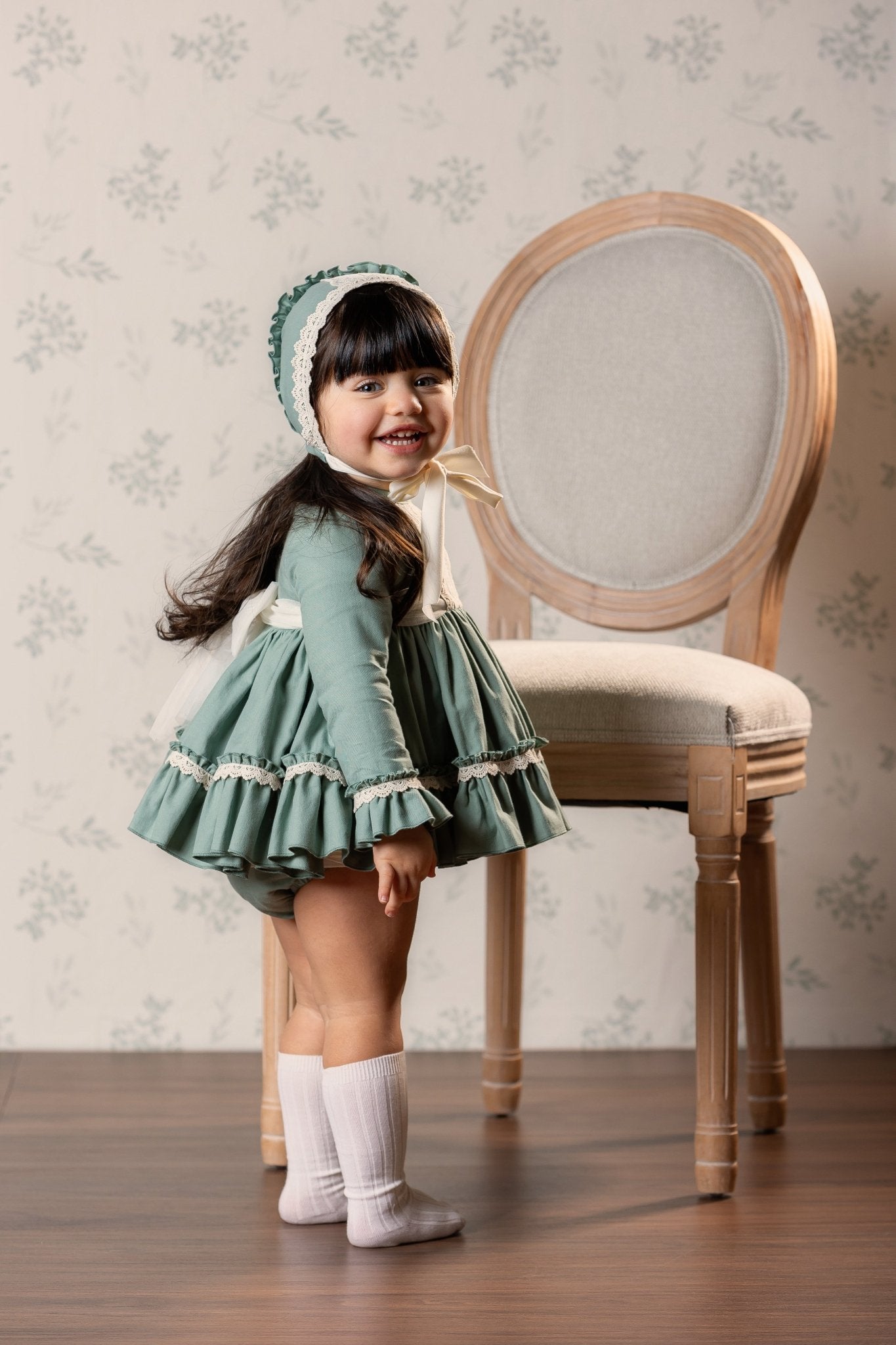 A young girl wearing a vintage-style sage green dress with Ivory lace trim, a matching bonnet, and cream socks, standing next to a wooden chair against a floral wallpaper background.