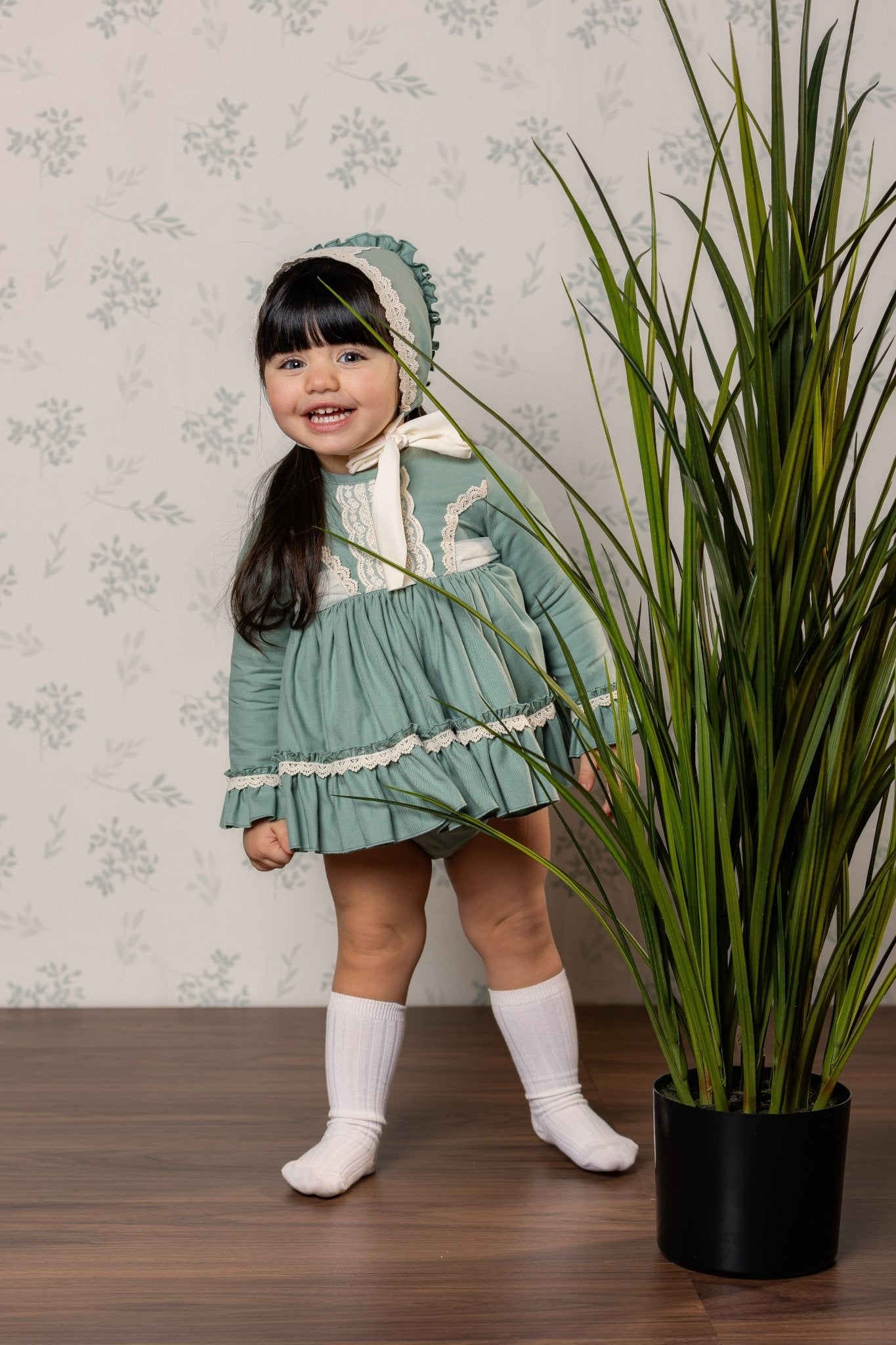 A young girl wearing a vintage-style sage green and ivory dress with a matching bonnet, cream knee-high socks, and standing on a wooden floor next to a potted green plant against a floral wallpaper background.