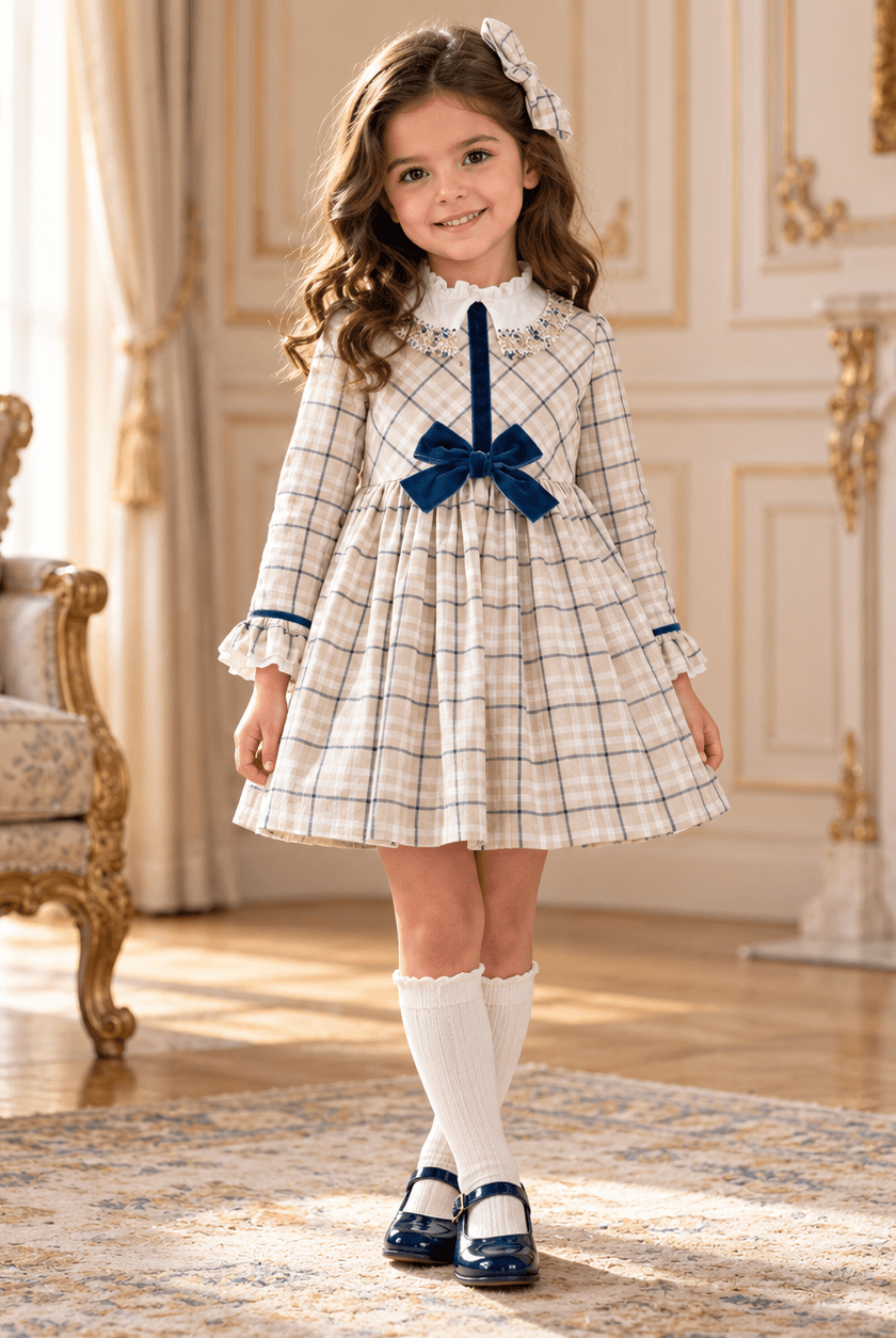 A young girl stands in an ornate, elegant room wearing a cream and navy plaid dress with a large navy bow at the waist, white knee-high socks, and navy Mary Jane shoes.