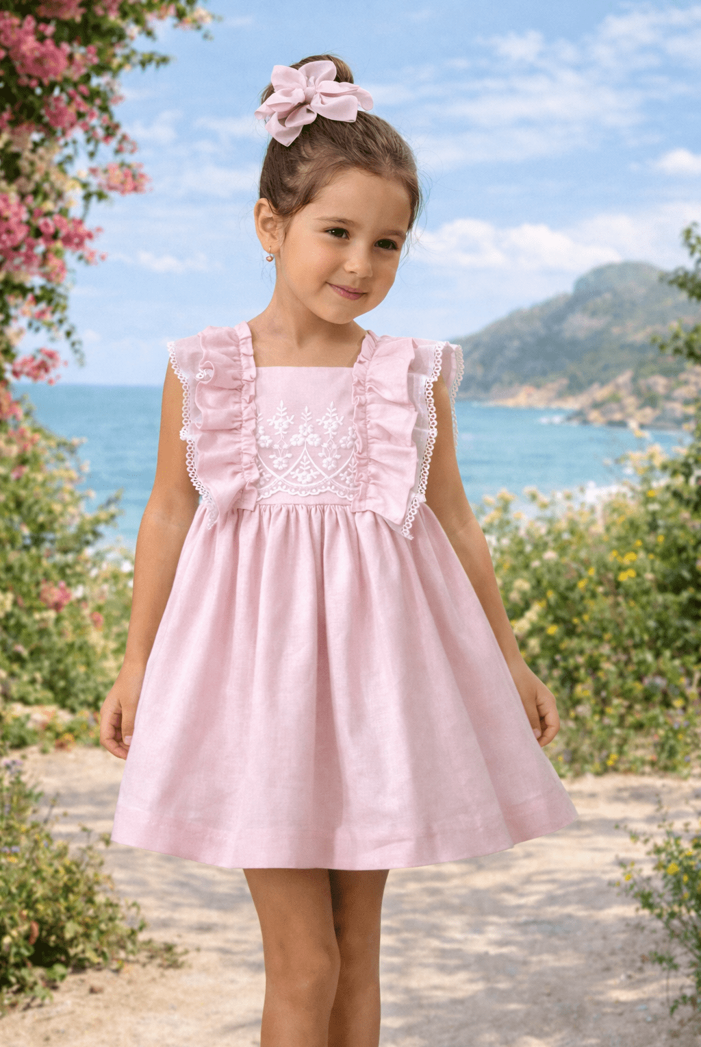 Young girl in a Basmarti SS26 pink dress with ruffled sleeves standing in a scenic outdoor setting with flowers and mountains.