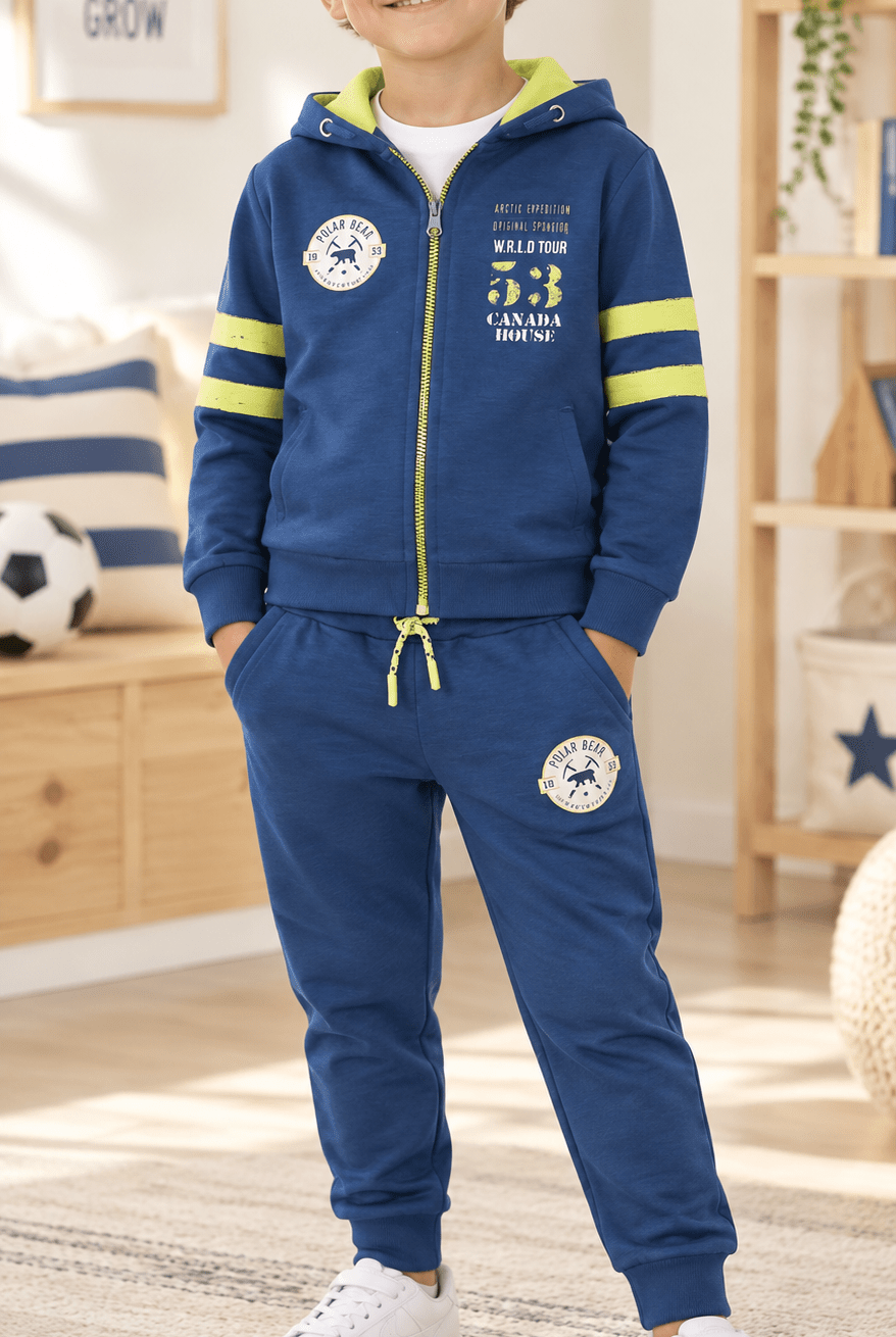 A young boy wearing a navy blue zip-up hoodie and matching jogger pants set with yellow-green accents, standing in a brightly lit child's bedroom.