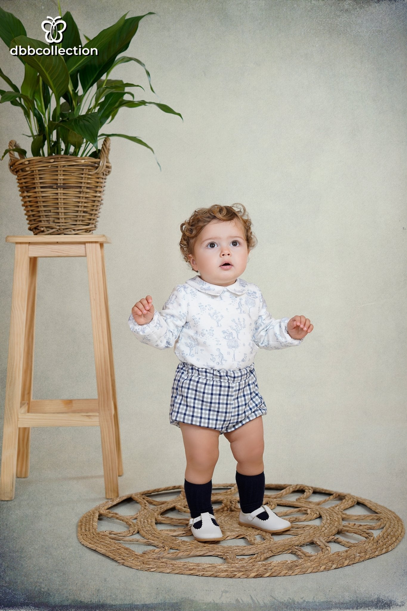 Baby boy wearing a blue woodland print shirt with Peter Pan collar and navy gingham shorts from DBB Collection AW25.