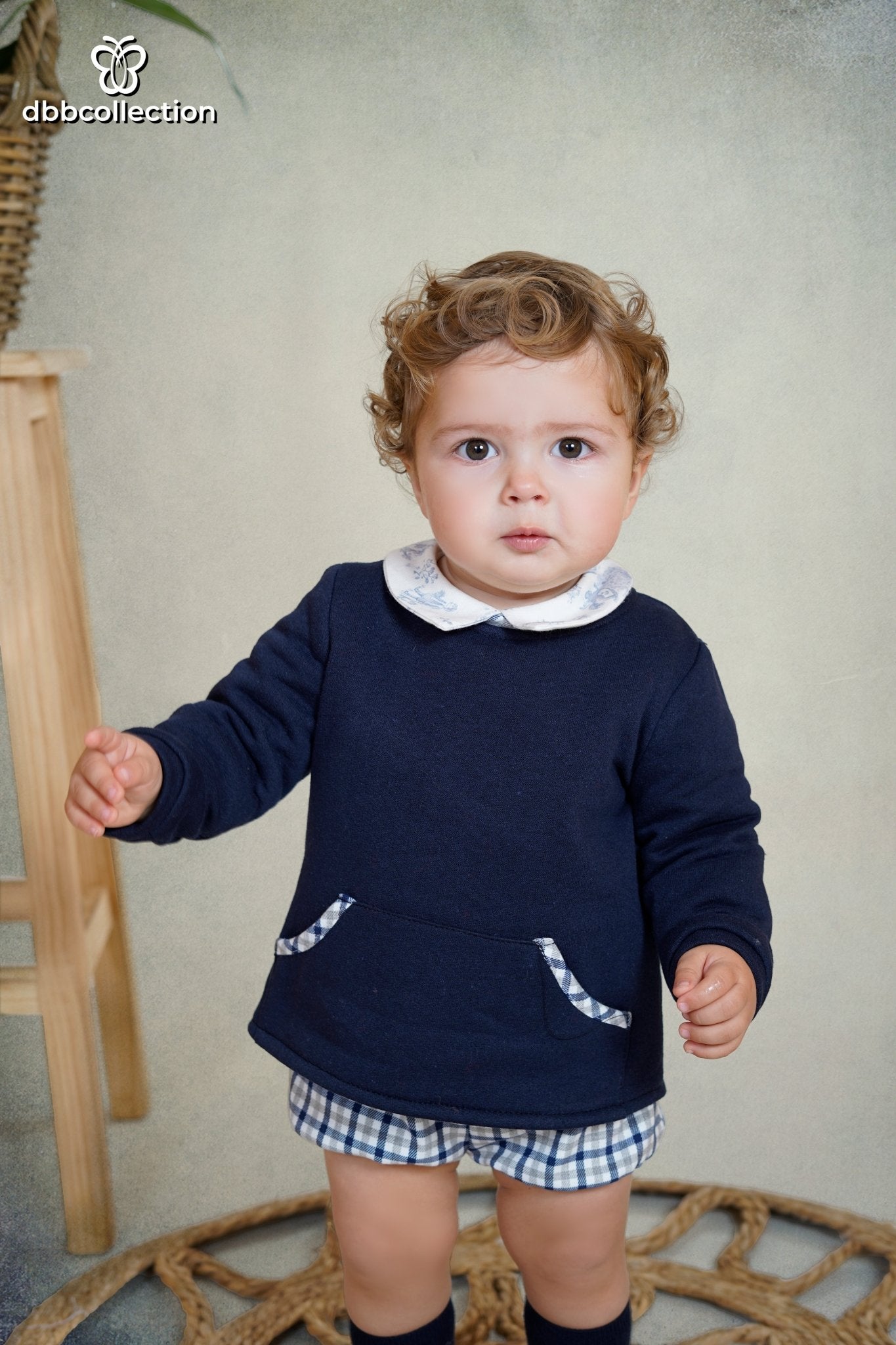 Baby boy wearing navy long-sleeve jumper with gingham trim pockets, over a printed shirt and matching gingham shorts.