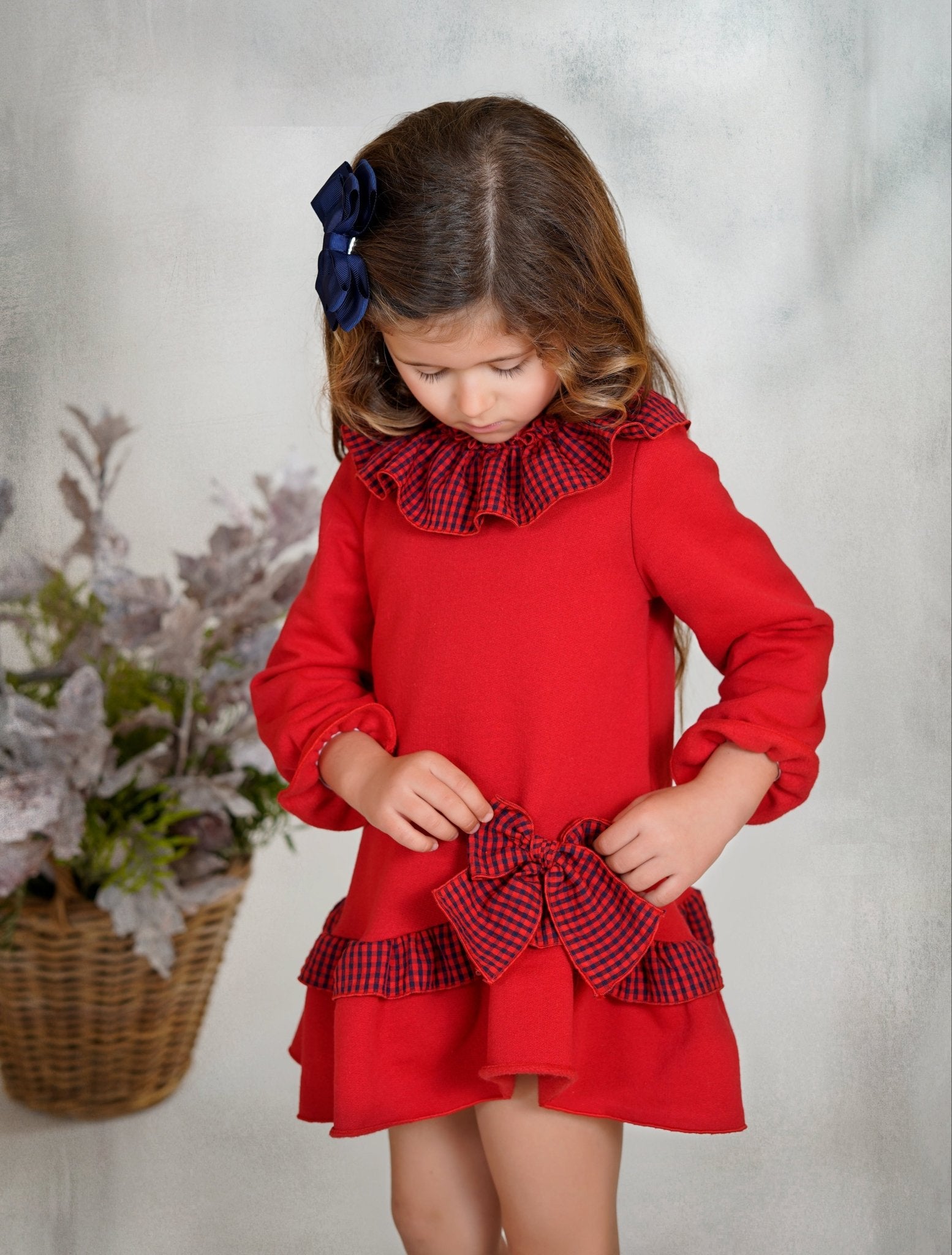 Girl wearing red long-sleeved holiday dress with navy gingham ruffle collar and bow, from DBB Collection AW25.