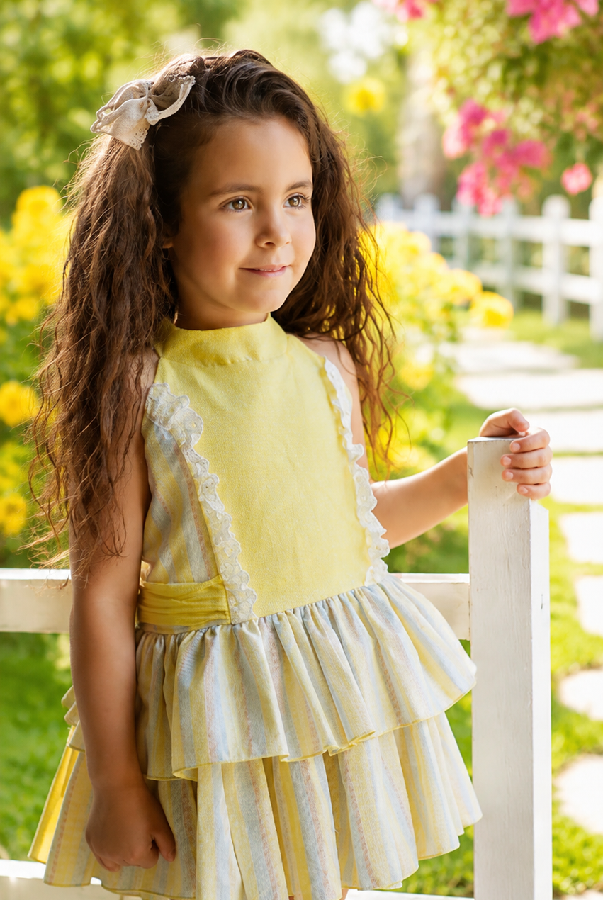 A young girl with long brown hair stands outdoors on a white fence in a sunny garden, wearing a yellow sleeveless dress with white lace trim and a tiered skirt.