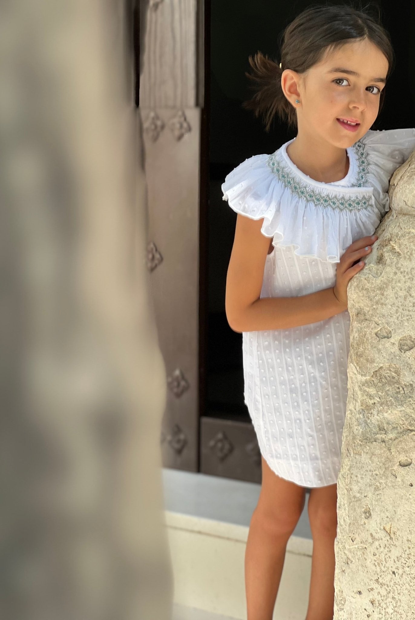 Young girl in a white smocked collar spanish dress standing against a stone wall by El Copo for SS26