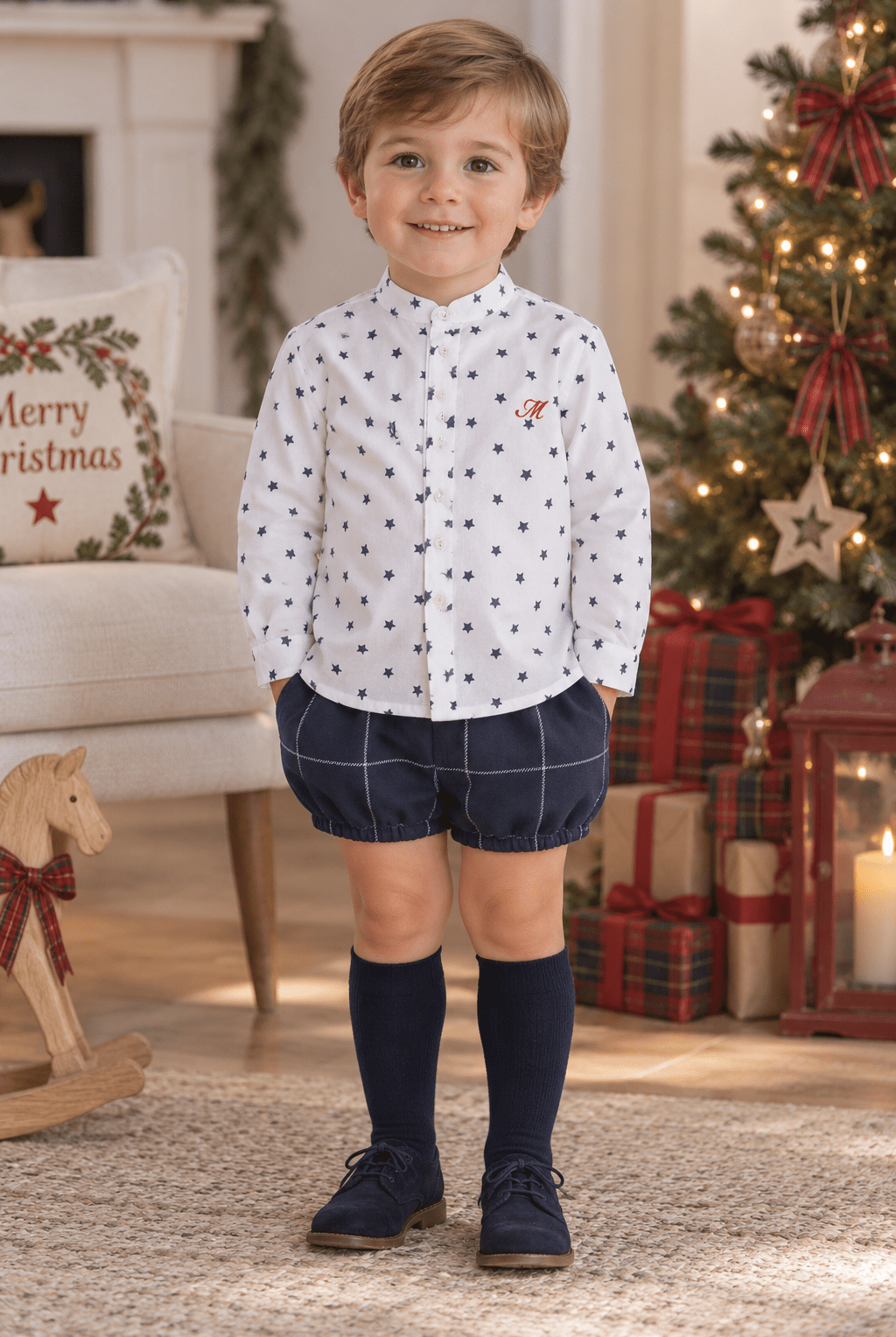 A young boy stands in a cozy holiday living room wearing a white long-sleeve button-up shirt with small navy polka dots, navy plaid shorts, navy knee-high socks, and navy shoes. A Christmas tree, wrapped gifts, and a fireplace are visible in the background.