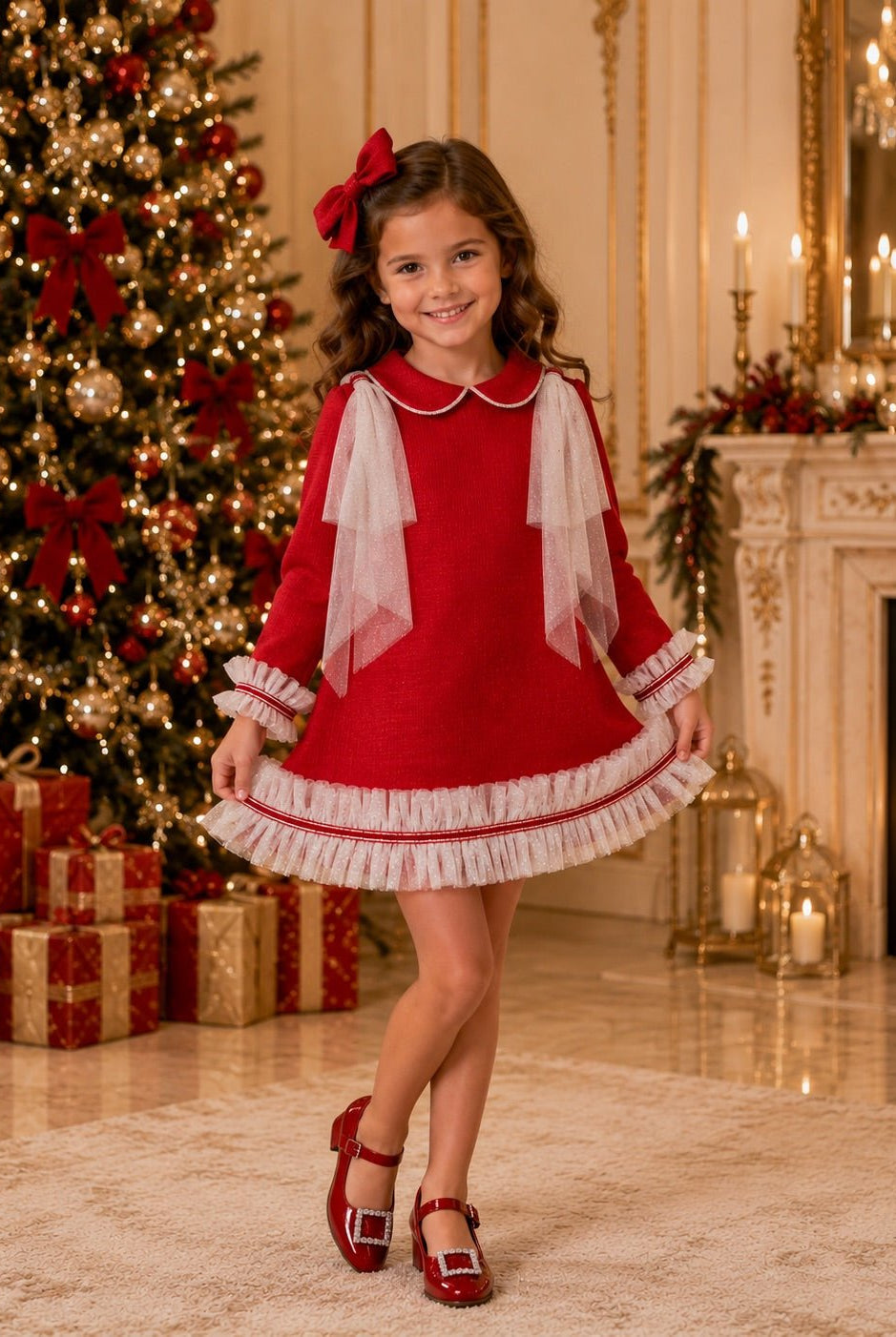 A young girl wearing a festive red and cream Naxos AW26 holiday dress with tulle cream bow accents, red shoes, and a red hair bow, standing in a decorated Christmas room with a tree, fireplace, and wrapped gifts.
