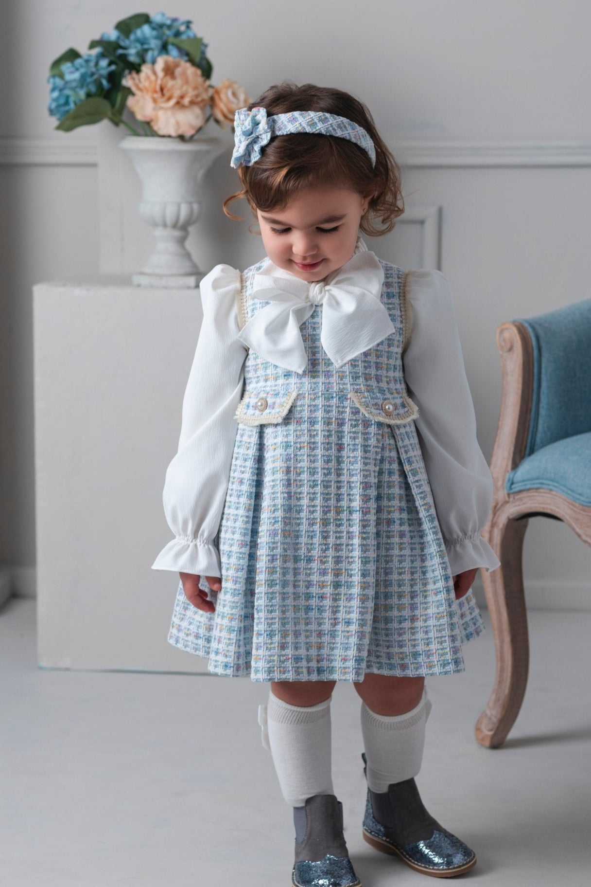 Young girl in a checkered dress with a white blouse and headband, standing indoors.