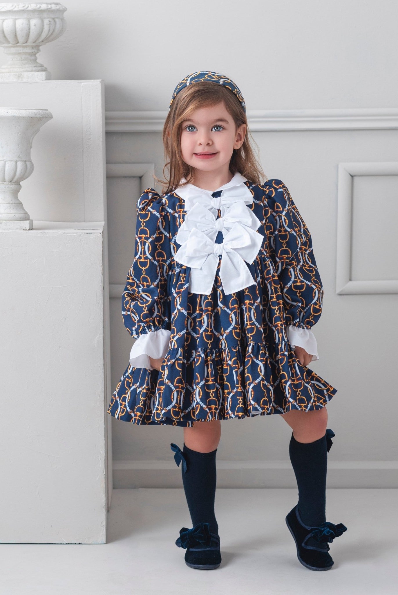 Young girl in a patterned Rochy AW25 Vintage Chains Navy and Gold dress with white bows standing in a room with decorative elements.