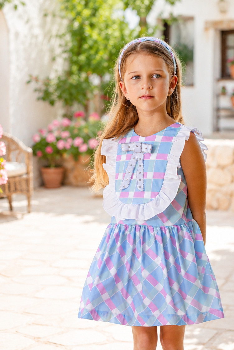 A young girl stands in a sunlit outdoor courtyard wearing a sleeveless blue and pink plaid dress with a white ruffled collar and bow detail. Pink flowers and greenery frame the scene.