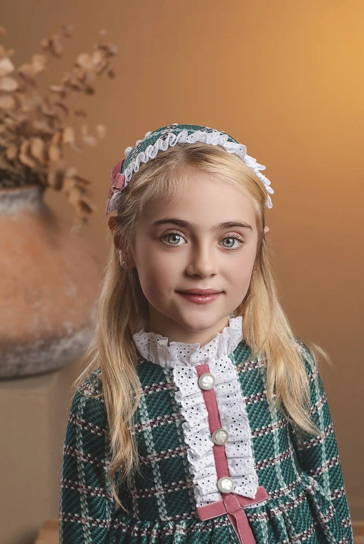 A young girl with blonde hair stands wearing a green and white plaid dress with a pink bow and white lace collar, paired with a matching headband. She is posed in a warm, rustic indoor setting with a wooden bench and a large terracotta vase in the background.