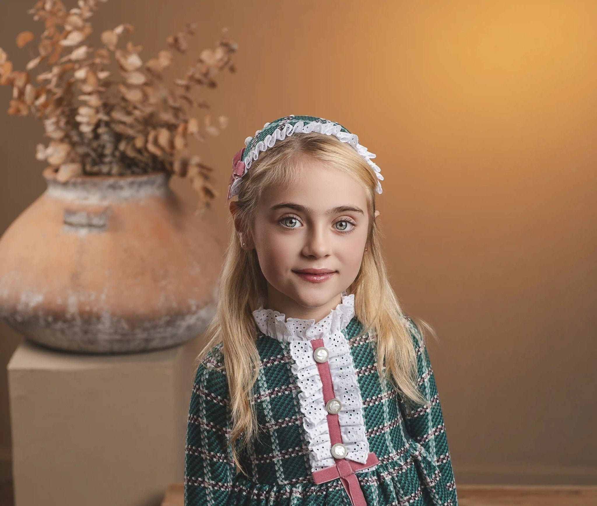 A young girl with blonde hair stands wearing a green and white plaid dress with a pink bow and white lace collar, paired with a matching headband. She is posed in a warm, rustic indoor setting with a wooden bench and a large terracotta vase in the background.