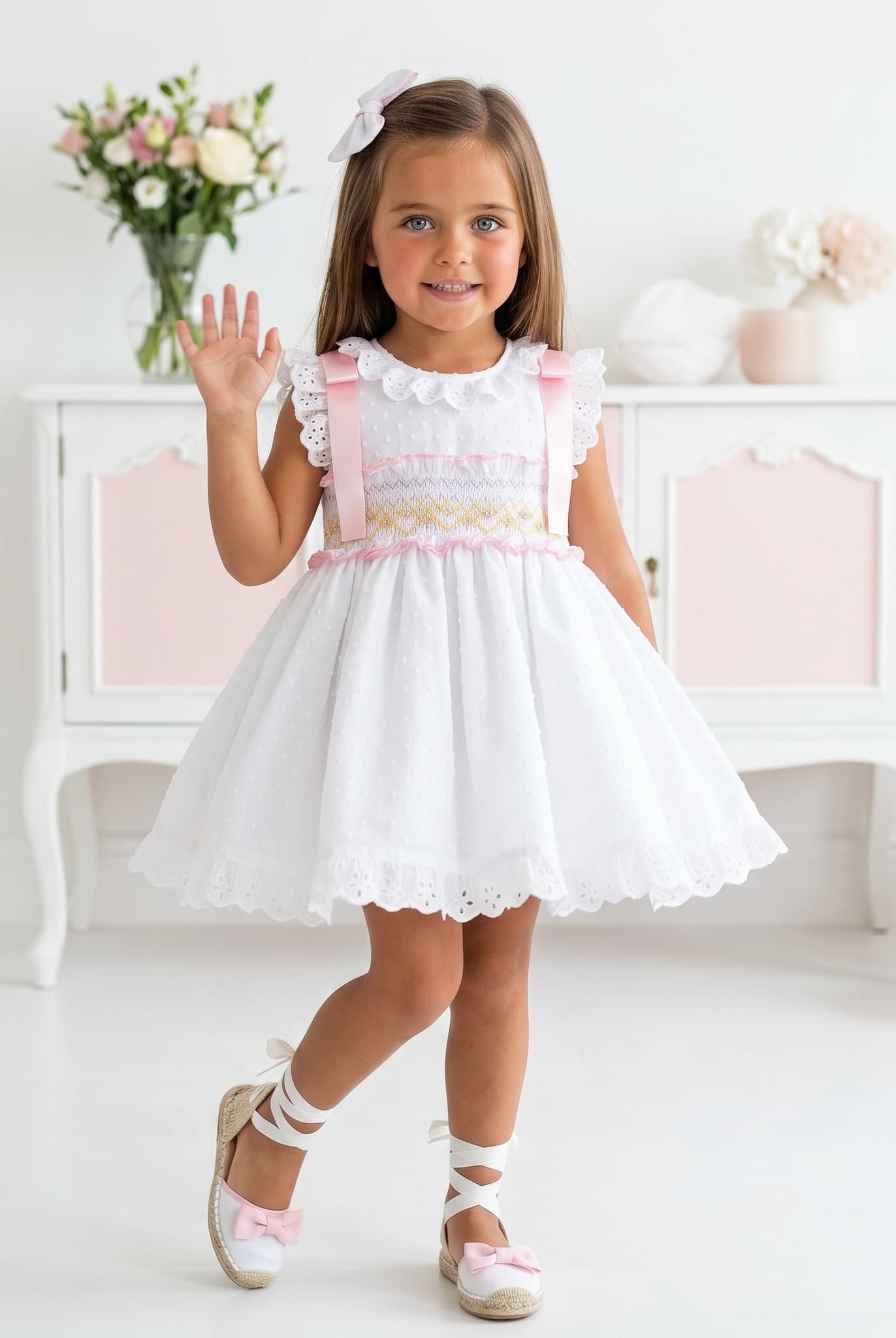 Young girl in a Sonata white smocked dress with pink accents waving in a bright room.