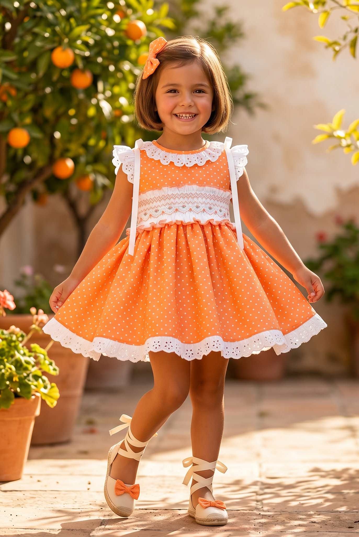 Young girl in an Sonata orange smocked dress with white lace trim standing in a sunlit outdoor setting with plants and fruit trees.