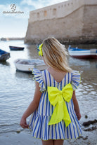 A young girl with blonde hair is seen from behind, wearing a blue and white striped dress with a large neon yellow bow on the back and a matching yellow hair clip, standing on a beach with water and a stone fort in the background.