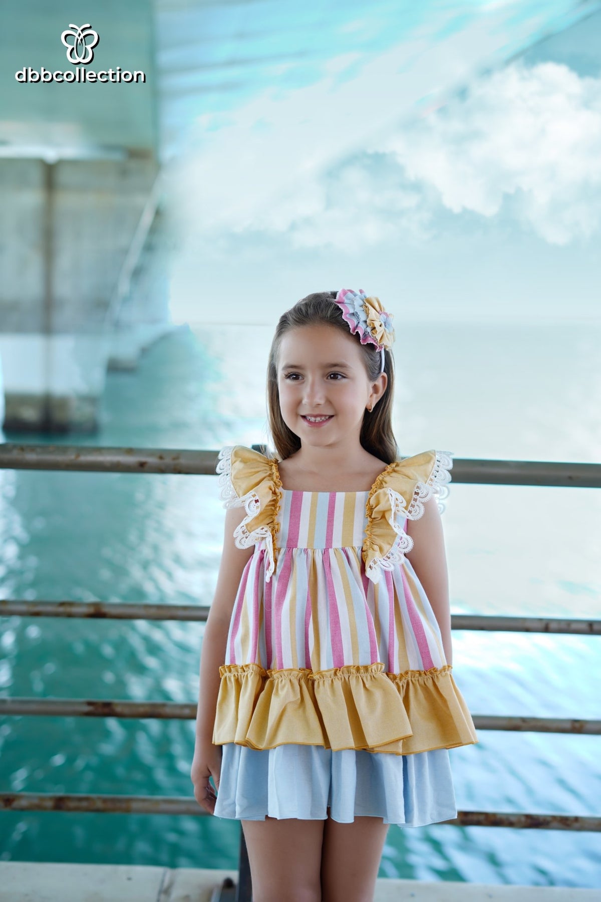 A young girl with a floral hair accessory stands in front of a railing, wearing a sleeveless ruffled top with pastel vertical stripes and a yellow ruffled hem, paired with a light blue skirt.