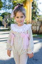 A young girl wearing a Naxos AW26 pink pastel multicolor knit romper with a large white ruffled collar, pink bows, and a pink hair bow, standing outdoors on a paved path.