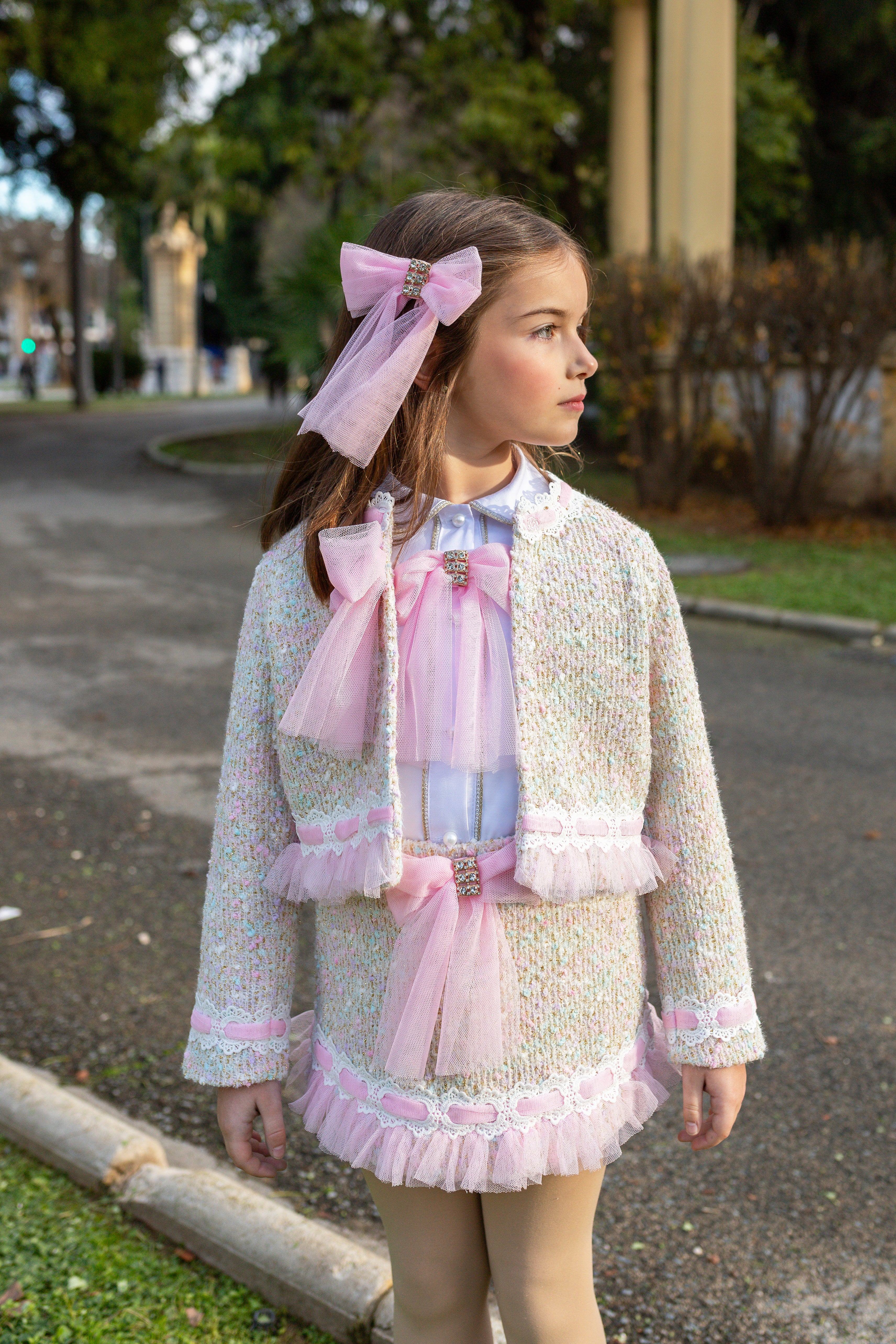 A young girl wearing a Naxos AW26 pastel pink and cream tweed jacket with matching ruffled trim, a large pink bow headband, and a matching pink bow at the waist, standing outdoors on a paved path.