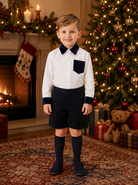 A young boy stands in a holiday-themed room wearing a white long-sleeve shirt with dark navy collar, cuffs, and chest pocket, paired with navy shorts and navy knee-high socks. A decorated Christmas tree, fireplace, and gifts are visible in the background.