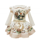 A long-sleeve baby dress in cream with a detailed woodland animal and floral print on the bodice and skirt, featuring a large satin bow at the waist and a decorative collar.