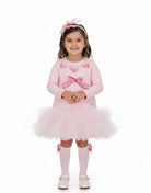 A young girl stands against a plain white studio background wearing a light pink long-sleeve dress with a tulle tutu skirt, pink bow details, matching pink socks, and a pink bow headband.