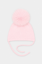 A light pink knit baby beanie hat with a fluffy pom-pom on top and matching tie strings, shown against a plain light gray background.