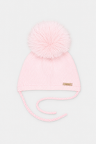A light pink knit baby beanie with a large fluffy pom-pom on top and matching ear ties, shown against a plain light gray background.