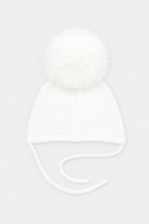 A white knit baby beanie hat featuring a large fluffy pom-pom on top and matching white ear tie strings, shown against a light gray background.
