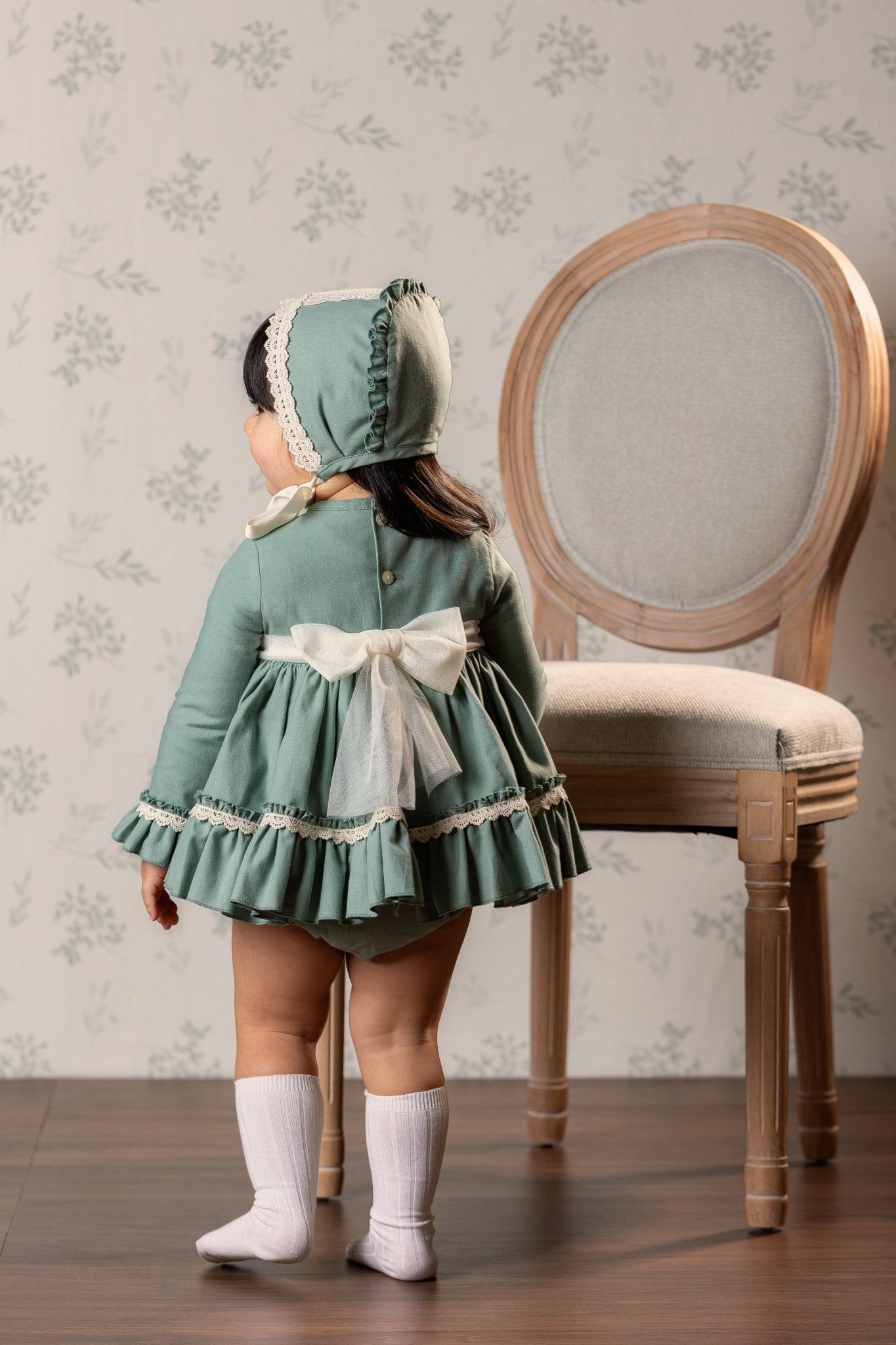 A toddler girl viewed from behind wearing a sage green long-sleeve dress with a Ivory bow and bonnet, standing next to a wooden chair in front of floral wallpaper.