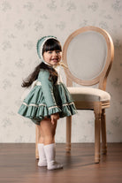 A young girl wearing a vintage-style sage green dress with Ivory lace trim, a matching bonnet, and cream socks, standing next to a wooden chair against a floral wallpaper background.