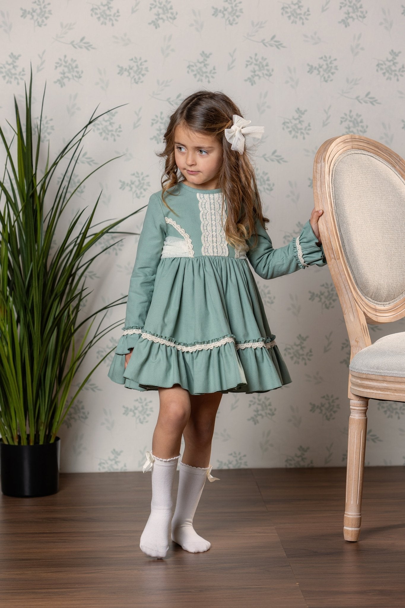 A young girl with long brown hair wearing a dusty green long-sleeve dress with cream lace details and cream knee-high socks, standing in a room with patterned wallpaper, a wooden chair, and a potted plant.