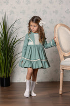 A young girl with long brown hair wearing a dusty green long-sleeve dress with cream lace details and cream knee-high socks, standing in a room with patterned wallpaper, a wooden chair, and a potted plant.