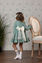 A young girl viewed from behind wearing a sage green long-sleeve dress with a large cream bow at the back, cream socks, and  hair bow, standing in a room next to a wooden chair against a floral wallpaper background.