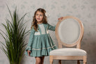A young girl with long light brown hair wearing a sage green long-sleeve dress with ivory lace trim stands next to a light wood and upholstered chair, holding its backrest against a patterned wallpaper background with a potted plant on the left.