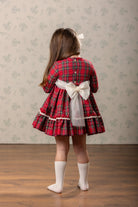 A young girl viewed from behind wearing a red tartan plaid dress with a large ivory  bow at the back, cream knee-high socks, and a white hair bow, standing on a wooden floor against a patterned wallpaper background.