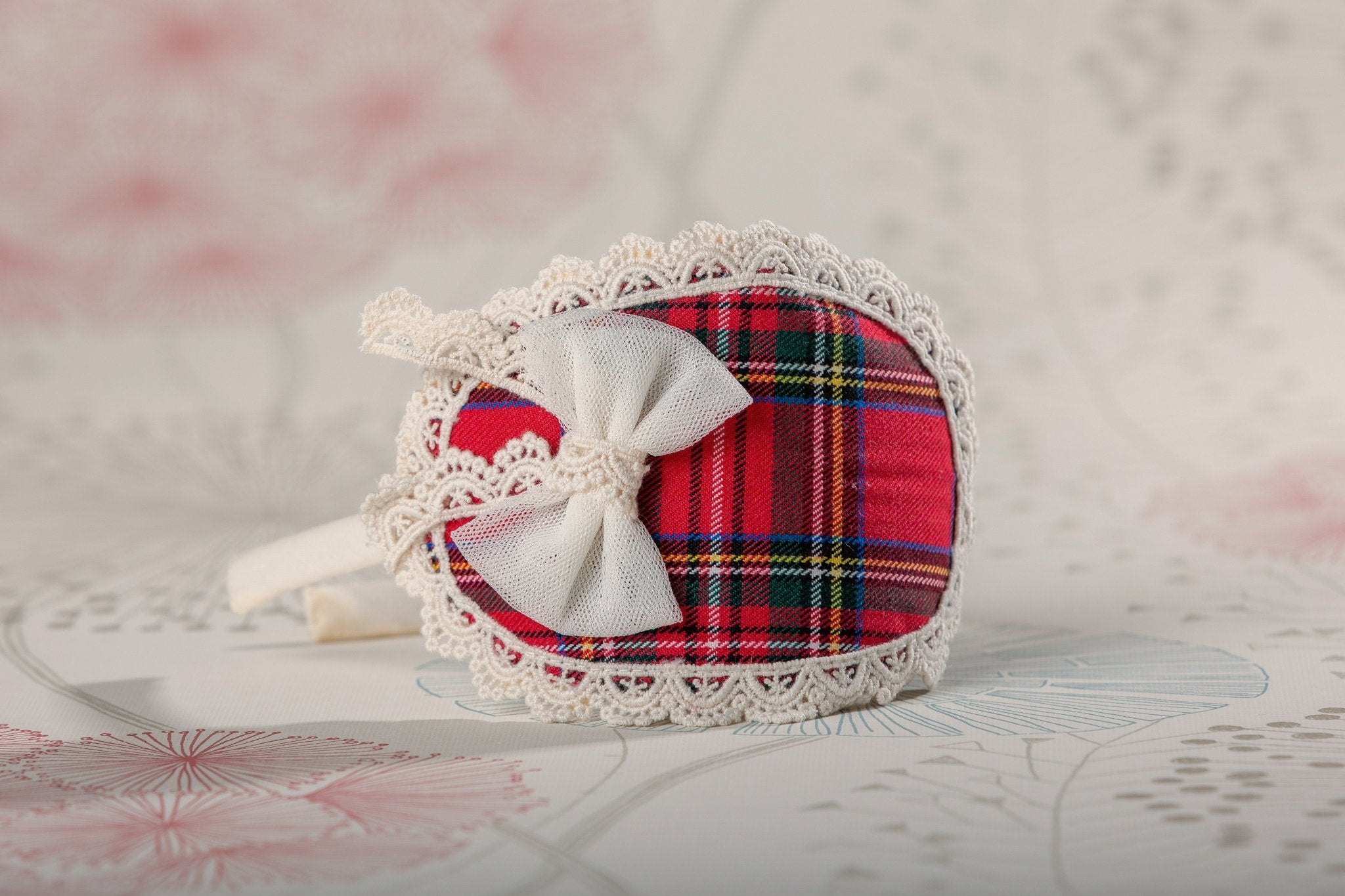A small decorative accessory with a red tartan plaid fabric body, white lace trim, and a white bow, photographed on a soft light background.