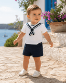 A toddler boy wearing a white short-sleeve sailor shirt with navy blue trim and a matching navy blue shorts, standing outdoors on a stone path near a blue window and potted flowers.