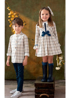 A boy and a girl modeling coordinated plaid outfits in front of a textured olive-green backdrop. The boy wears a beige and white plaid long-sleeve shirt with dark blue pants and white sneakers.