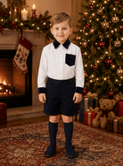 A young boy stands in a holiday-themed room wearing a white long-sleeve shirt with dark navy collar, cuffs, and chest pocket, paired with navy shorts and navy knee-high socks. A decorated Christmas tree, fireplace, and gifts are visible in the background.