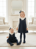 Two young girls in matching navy blue boucle pinafore dress and matching romper with ivory ruffled long-sleeve shirts, standing and sitting together in a bright, elegant room with cream-colored furniture.