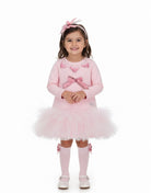 A young girl stands against a plain white studio background wearing a light pink long-sleeve dress with a tulle tutu skirt, pink bow details, matching pink socks, and a pink bow headband.