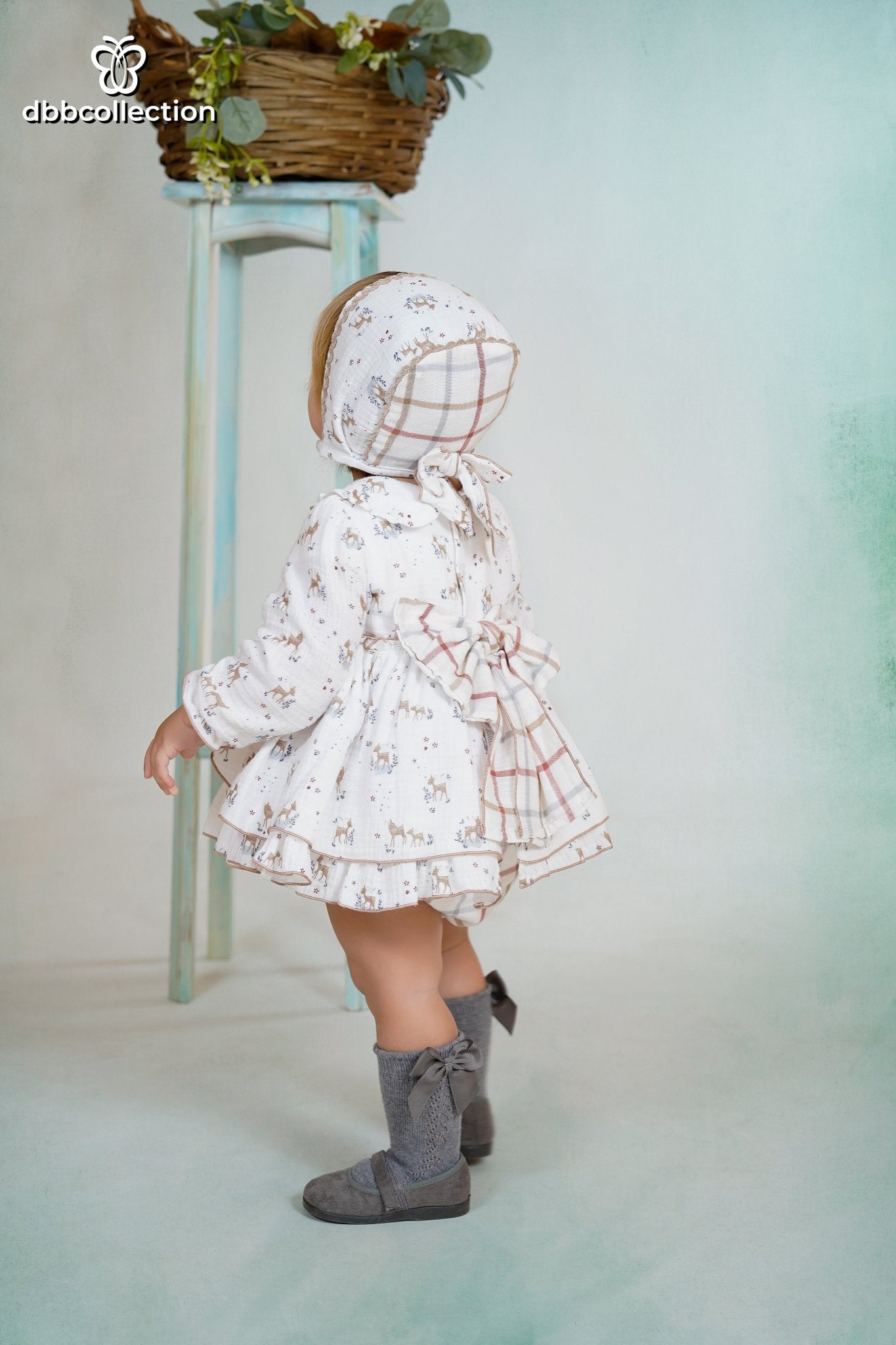 Baby wearing a white dress with deer print, matching bonnet, and ruffle details from DBB Collection AW25.